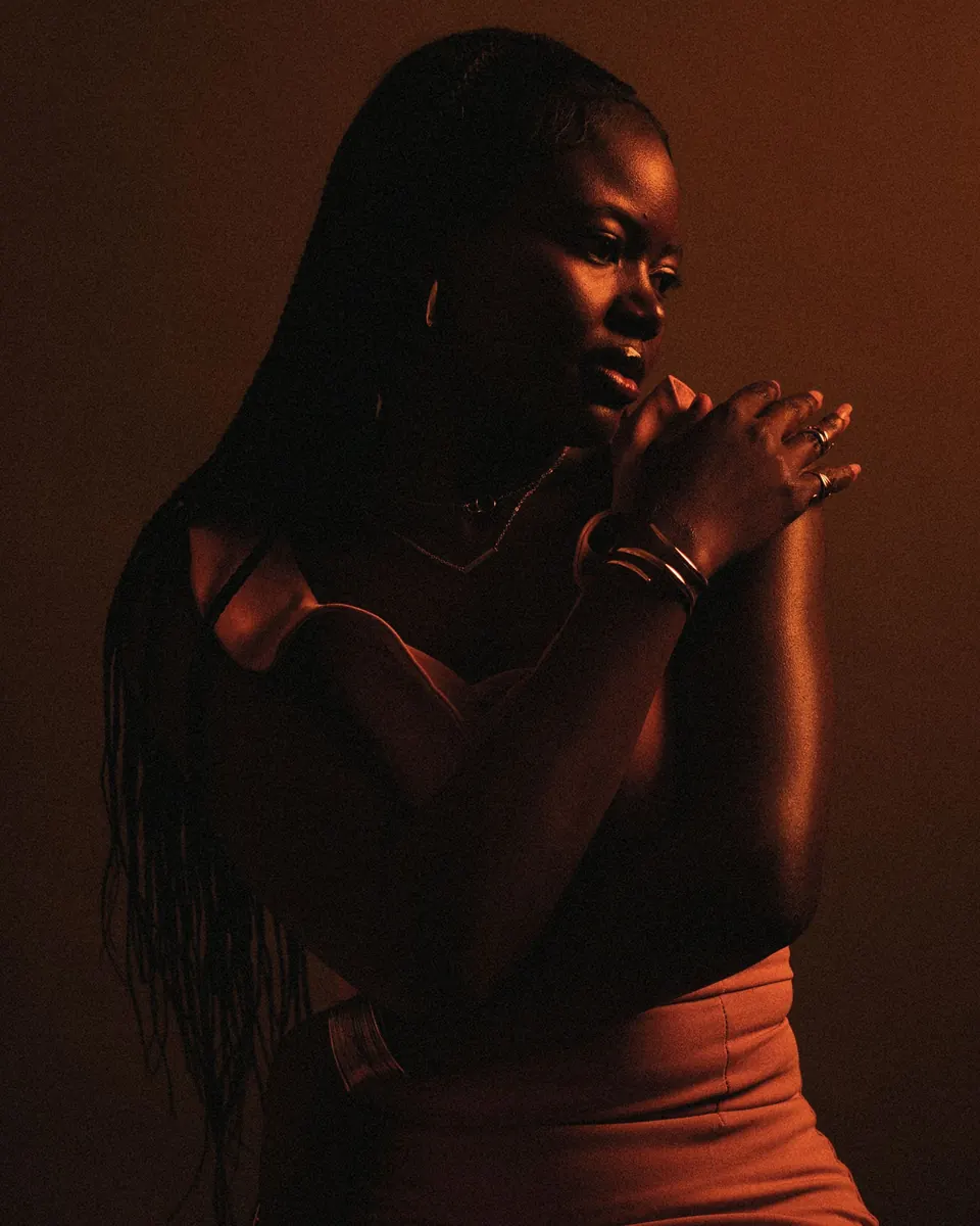 Woman with long braids in warm amber light, hand resting at her collarbone
